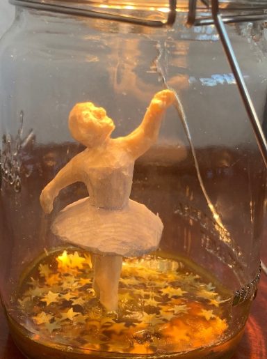 Eine ballerinaartige Figur in einem Glasbehälter, umgeben von goldenen Sternen.